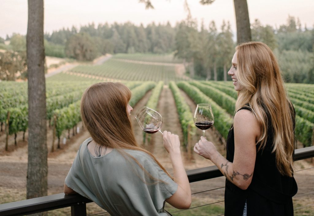 Two women standing on a balcony drinking holding wine glasses while looking out at the vineyard.